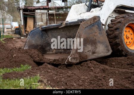 Un petit véhicule de chantier avec une roue orange et un chargeur frontal déplace le sol près d'une structure rustique avec un toit en métal ondulé et des panneaux en bois. Banque D'Images