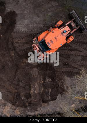 Une vue aérienne d'un tracteur orange sur un sol labouré foncé, montrant des traces de pneus circulaires, des accessoires et une végétation clairsemée dans le coin. Banque D'Images