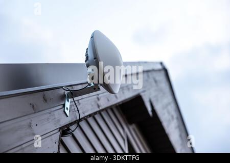 Un routeur Wi Fi en forme de dôme blanc est monté sur le bord du toit d'une maison en bois. Les câbles sont soigneusement fixés, avec un ciel couvert en arrière-plan. Banque D'Images