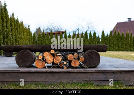 Banc en bois rustique avec du bois de chauffage en dessous sur une plate-forme, bordé par de grands arbres à feuilles persistantes. Les toits voisins et une cheminée sont visibles dans le backgro Banque D'Images