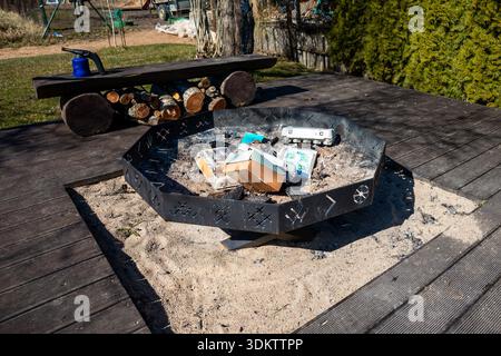 Un foyer hexagonal en métal rempli d'objets jetés sur une base sablonneuse, entouré d'une terrasse en bois, d'un banc rustique et d'éléments extérieurs. Banque D'Images