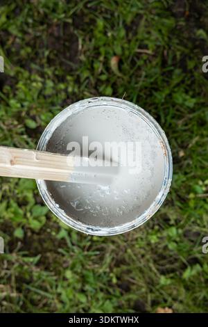 Une boîte de peinture gris clair avec un bâtonnet en bois est vue d'en haut, placé sur l'herbe avec des taches de sol. La surface peinte apparaît lisse et pétillante. Banque D'Images