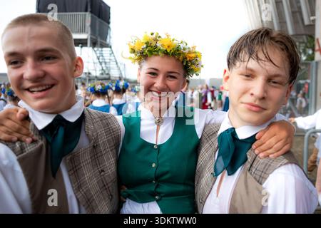 Trois enfants en costumes nationaux lettons, avec une fille en couronne florale et des garçons en gilets à carreaux, debout ensemble avec une foule et une structure métallique Banque D'Images