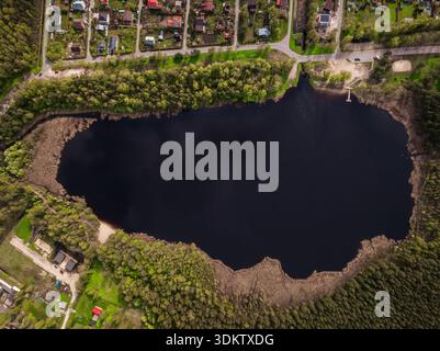 Une vue aérienne d'un lac sombre avec un quai sur la droite, entouré d'arbres et d'un quartier résidentiel avec des toits colorés et des routes planifiées. Banque D'Images
