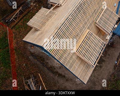 La vue aérienne montre un toit à pignon abrupt en construction avec des lattes, une sous-couche sombre, des ouvertures de lucarnes encadrées, des feuilles tombées, des échelles, lumbe empilé Banque D'Images