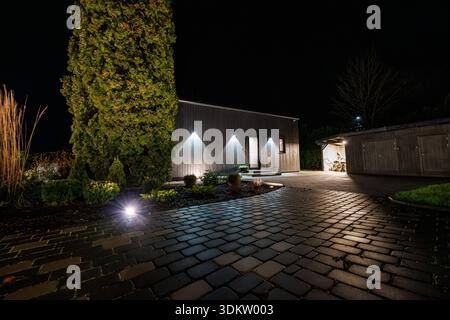 Une maison basse en bois projette des bassins de lumière sur des pavés humides tandis qu'un grand conifère est mis en lumière. Du bois de chauffage et une dépendance en bois se trouvent à proximité, capturés Banque D'Images