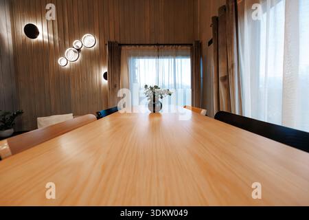Une longue table en bois pâle mène à un vase noir avec de l'eucalyptus, des rideaux transparents, de hautes fenêtres et des panneaux de bois verticaux. Les appliques murales circulaires brillent Banque D'Images