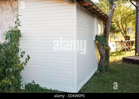 Un petit hangar en bois blanc avec un toit incliné et des volets se dresse près d'un arbre penché, entouré d'herbe, de buissons et d'une table en bois avec des chaises. Banque D'Images