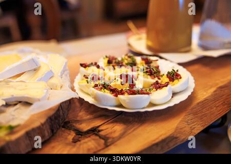 Des œufs dévorés en deux sont assis sur une assiette blanche avec du bacon et des microverts sur une planche de bois à côté de fromage à croûte molle dans une lumière chaude du soir, stylisée pour Banque D'Images