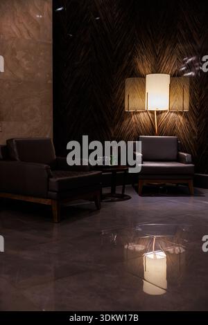 Fauteuils modernes en cuir avec carafe par mur en bois à chevrons apparaît dans un coin salon. Un lampadaire projette une lumière chaude et des reflets nets sur le vernis Banque D'Images