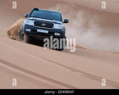 Vue de face d'une Toyota Land Cruiser noire alimentant une dune de sable doré abrupte, des roues dégageant des nuages de sable dans le vaste désert du Sahara marocain. Banque D'Images