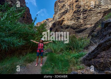 Randonneur féminin avec les bras tendus sautant sur le sentier de la gorge de Masca entouré d'une végétation luxuriante et de falaises imposantes Banque D'Images