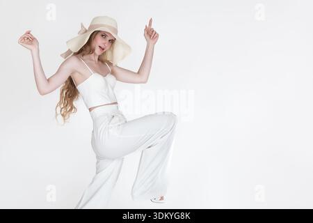 Femme à la mode dans une tenue élégante tout blanc et chapeau d'été frappe la pose ludique. Photo studio haute clé, pointant vers le haut sur fond blanc. AMPL Banque D'Images