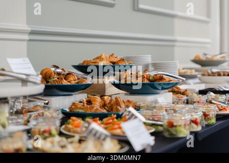 Buffet de restauration élégant avec croissants frais, canapés savoureux, salades dans des bocaux en verre et assortiment d'amuse-gueules présentés sur des stands étagés pour les entreprises Banque D'Images