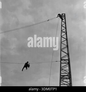Sloviansk, région de Donetsk, Ukraine, URSS. 1960s. Une photographie d'archives dramatique à bas angle de deux hommes de ligne effectuant des travaux dangereux à haute altitude sur une ligne de transmission d'énergie. Un ouvrier est perché au sommet d'une tour en treillis métallique, tandis que son collègue est suspendu aux fils en l'air contre un ciel nuageux, installant des isolateurs. Cette image authentique en noir et blanc capture la bravoure, les risques et les compétences techniques des travailleurs assurant l'électrification de la région industrielle pendant l'ère soviétique stable et pacifique. Banque D'Images