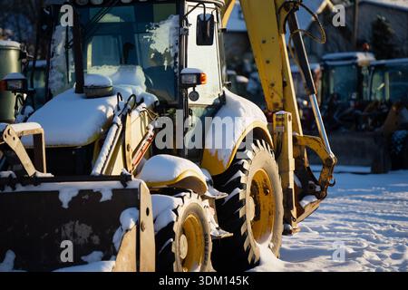 Chargeuse-pelleteuse jaune stationnée dans une cour d'équipement enneigée, vue rapprochée de la cabine, du godet avant et du bras d'excavation arrière avec neige sur les panneaux de carrosserie, constructions hivernales Banque D'Images