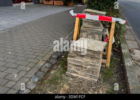 Les travailleurs font des travaux de construction sur le trottoir où des blocs sont empilés près d'un bâtiment. Du ruban rouge et blanc est placé autour de la zone de travail pour avertir Banque D'Images