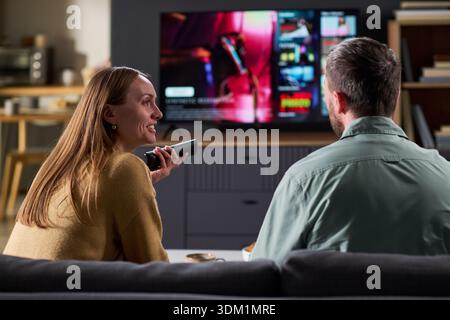 Femme jeune adulte caucasienne et homme jeune adulte caucasien assis sur le canapé regardant la télévision ensemble, femme tenant la télécommande et souriant tout en parlant à l'homme Banque D'Images