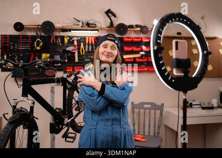 Souriant femme heureuse blogueuse influencer streaming en direct travaillant dans le garage, vlogger s'embrassant les yeux fermés sur la caméra pour examiner. mécanicien de vélo en s. Banque D'Images
