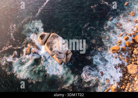 Vue aérienne des vagues mousseuses s'écrasant autour d'un énorme rocher alors que le rivage doré rencontre l'océan sombre et tourbillonnant, le Cap, Western Cape, South AF Banque D'Images