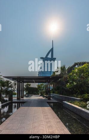 Dubaï, Émirats Arabes Unis - 28 octobre 2025 : vue imprenable sur l'hôtel Burj Al Arab à travers un sentier de jardin paysager avec lumière solaire Dubaï Banque D'Images