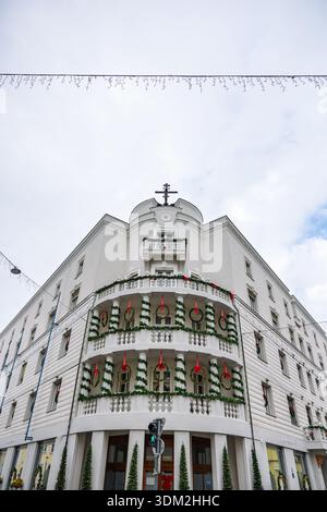 Un charmant bâtiment d'angle blanc drapé de guirlandes vertes et de rubans rouges, avec balcons et couronnes. Devantures de magasins au niveau de la rue et pavés enneigés Banque D'Images
