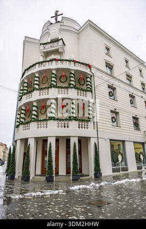Un charmant bâtiment d'angle blanc drapé de guirlandes vertes et de rubans rouges, avec balcons et couronnes. Devantures de magasins au niveau de la rue et pavés enneigés Banque D'Images