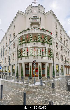 Un charmant bâtiment d'angle blanc drapé de guirlandes vertes et de rubans rouges, avec balcons et couronnes. Devantures de magasins au niveau de la rue et pavés enneigés Banque D'Images