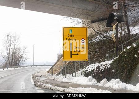 Un panneau routier jaune vif sous un passage supérieur dirige les voyageurs vers Celje, Teharje, Svetina et Lipa dans un cadre rural enneigé et pittoresque. Banque D'Images