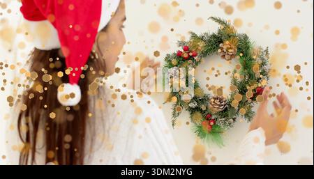 Ajustement femme indienne portant chapeau de Père Noël et pull blanc atteignant la couronne à la maison, avec des pommes de pin Banque D'Images