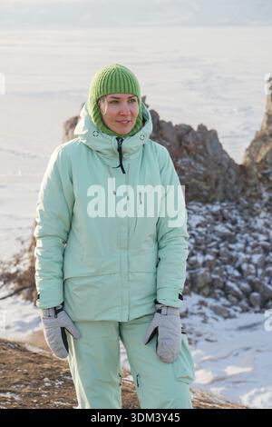 Jeune femme en vêtements chauds d'hiver se tient près de Shaman Rock dans le village de Khuzir sur l'île Olkhon, lac Baïkal, avec paysage glacé et rivage rocheux Banque D'Images