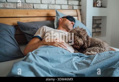 Chien Maltipoo au lit avec le propriétaire portant un masque pour les yeux de sommeil noir pendant la sieste de jour sur fond non focalisé. Animaux drôles et image de concept de style de vie sain Banque D'Images