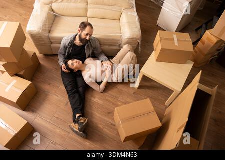 Vue de dessus du couple caucasien se relaxant sur le sol pendant la journée de déménagement, entouré de boîtes en carton dans le salon confortable. Heureux homme et femme se reposent tout en déballant dans le premier appartement propre. Banque D'Images