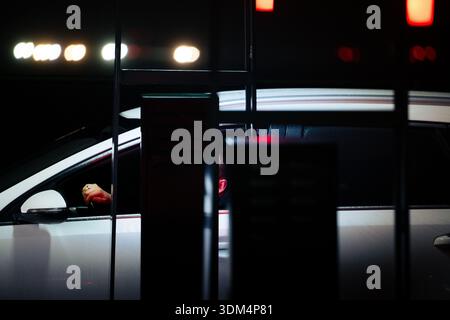 Un conducteur attend au point de commande d'un drive-in McDonald's la nuit, les mains sur le volant d'une voiture blanche sous des lumières artificielles brillantes du parking. Banque D'Images