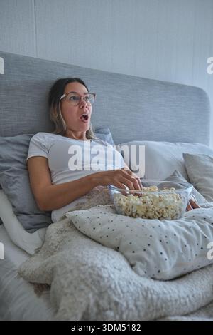 Femme se sentant surpris tout en se relaxant dans le lit manger du pop-corn et regarder un film Banque D'Images