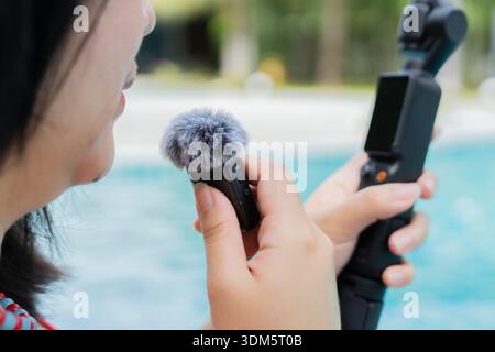 Gros plan jeune femme vlogger enregistrant une vidéo près d'une piscine et utilisant un microphone sans fil avec caméra de poche. Banque D'Images