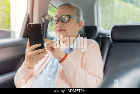 Femme asiatique senior utilisant son téléphone intelligent tout en étant assis sur le siège arrière dans la voiture moderne. Concept de vie de retraite. Banque D'Images