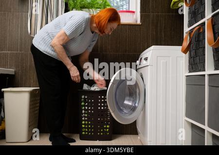Une femme mature met le linge dans la porte ouverte de sa machine à laver à chargement frontal. Elle est engagée dans des tâches quotidiennes d'entretien ménager à la maison. Banque D'Images