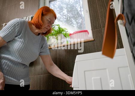 Vue de ver d'une femme de 70 ans qui lance un cycle de lavage en sélectionnant le programme approprié sur sa machine à laver domestique. Banque D'Images