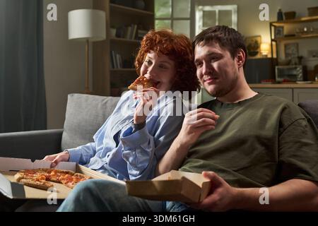 Jeune femme caucasienne et jeune homme caucasien assis sur un canapé mangeant de la pizza ensemble, souriant et paraissant détendu, tenant des tranches de pizza et des boîtes de pizza dans le cadre du salon Banque D'Images