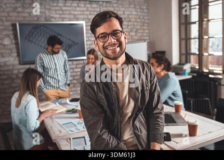 Portrait d'un jeune dirigeant masculin confiant debout et regardant la caméra tandis que ses collègues travaillent sur des conceptions architecturales en arrière-plan. Banque D'Images