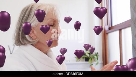 Femme senior debout portant un peignoir blanc dans la salle de bain, tenant la main droite, des coeurs violets, plante Banque D'Images
