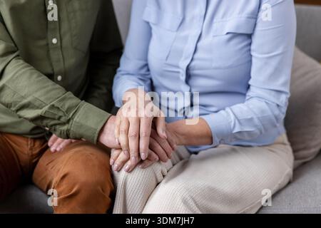 Homme mûr plaçant doucement sa main sur la main d'une femme plus âgée, transmettant le soin, l'empathie et une connexion forte entre eux tout en étant assis ensemble sur un canapé Banque D'Images