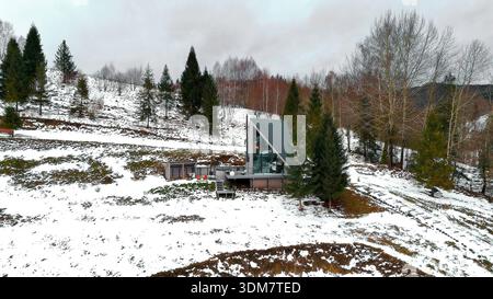 Maison triangulaire moderne dans un cadre enneigé à flanc de colline, maison à ossature en a avec de grandes fenêtres nichée sur une colline enneigée avec des arbres. Banque D'Images