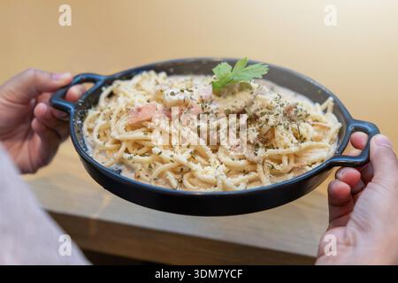 Mains tenant une poêle noire remplie de spaghetti carbonara crémeux, garnie de jambon de poulet, de parmesan et de persil frais. Banque D'Images