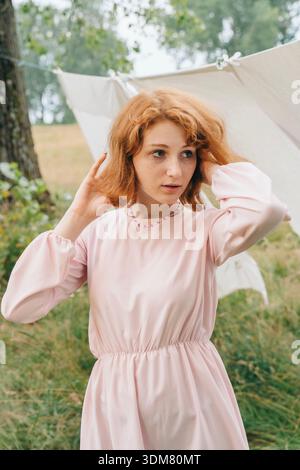 Jeune femme aux cheveux roux portant une robe rose douce debout à l'extérieur près des draps de lin séchant. Lumière naturelle, cadre de campagne, calme et serein atmos Banque D'Images