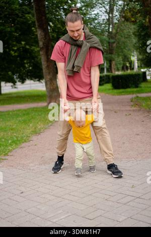 Un tout-petit heureux dans une chemise jaune vif fait ses premiers pas avec les mains de soutien de son père dans un parc verdoyant, capturant un moment d'apprentissage, de confiance et de liens familiaux. Banque D'Images