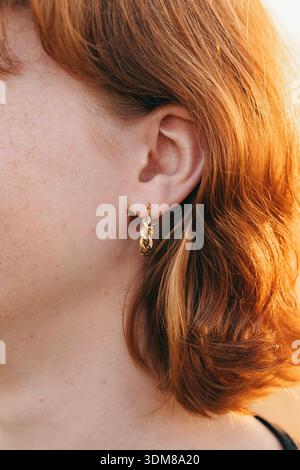 Photo en gros plan d'une femme aux cheveux roux portant une petite boucle d'oreille en or torsadée. L’image met en valeur la texture et la brillance de la boucle d’oreille par rapport au modèle Banque D'Images