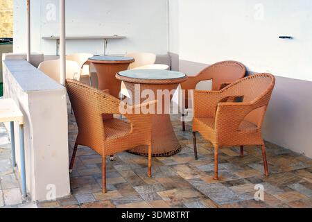 Café extérieur méditerranéen à budget simple en bord de route avec des chaises et des tables en résine tissée brune usée sur un patio carrelé avec une cloison en béton blanc. Banque D'Images