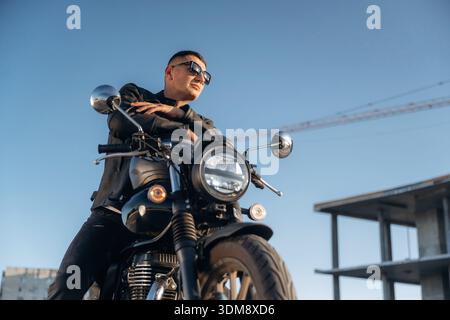 Contre le ciel dégagé, en lunettes de soleil. L'homme est dehors avec sa moto. Banque D'Images